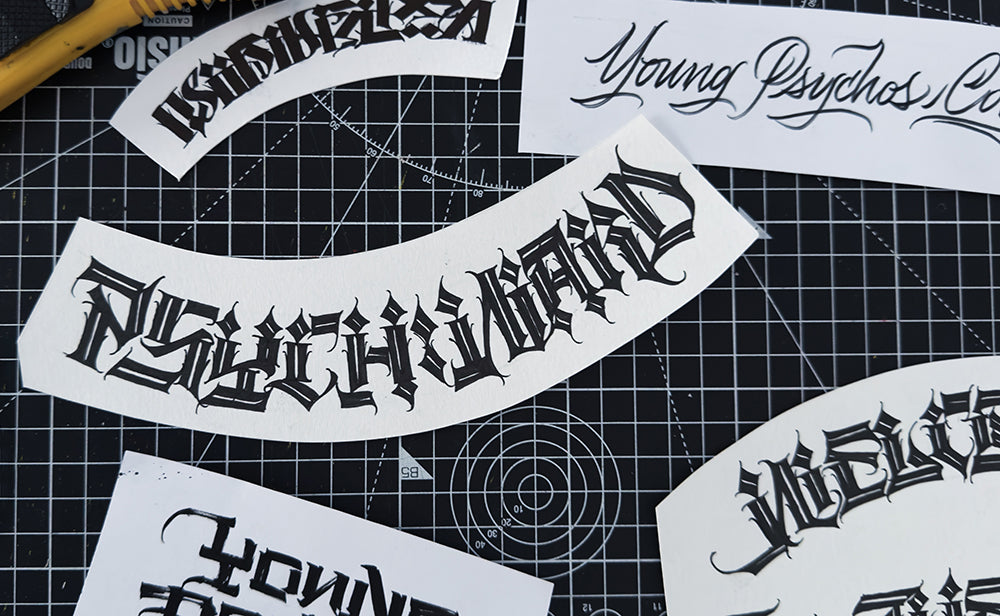Black lettering designs on white paper sheets on a black cutting mat with grid lines.