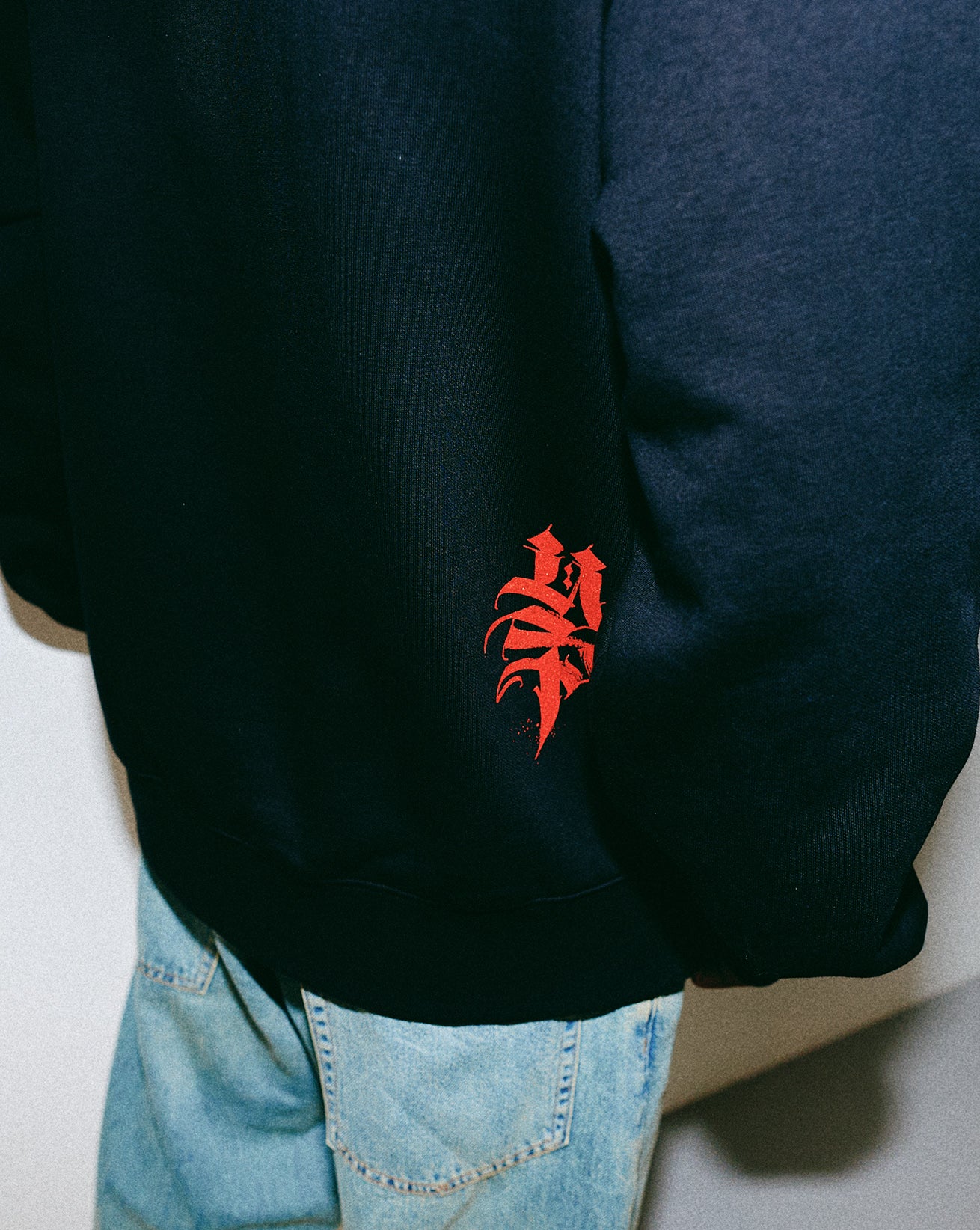 Black hoodie with red logo worn by a person, with light blue jeans visible.