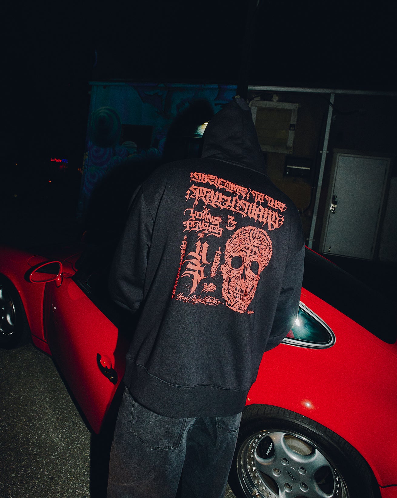 Person wearing a black hoodie with red graphics in front of a red car at night.