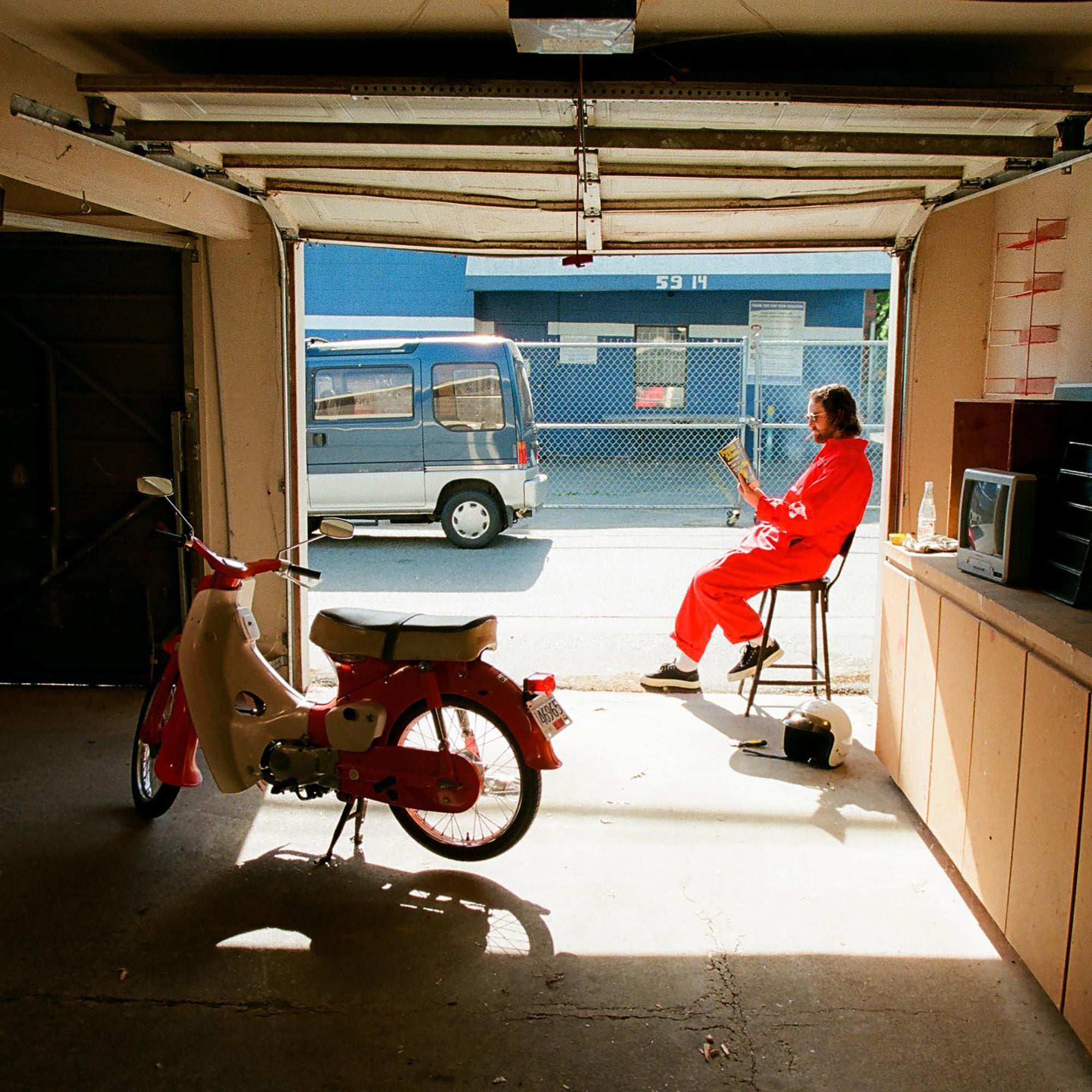 Person in red outfit sitting on a stool in a garage with a red scooter and a blue van outside.