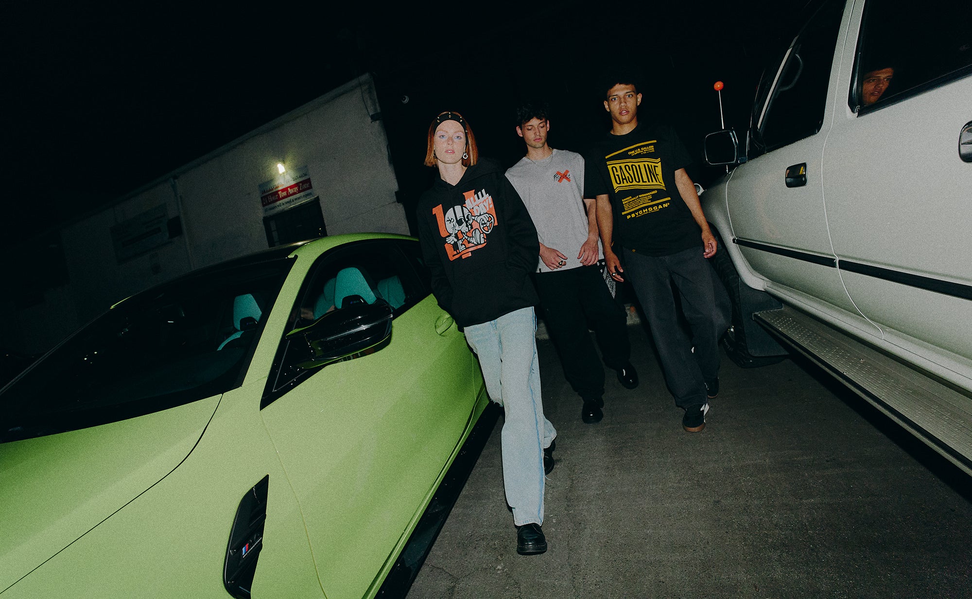 Three individuals walking near a lime green car and a white truck at night.