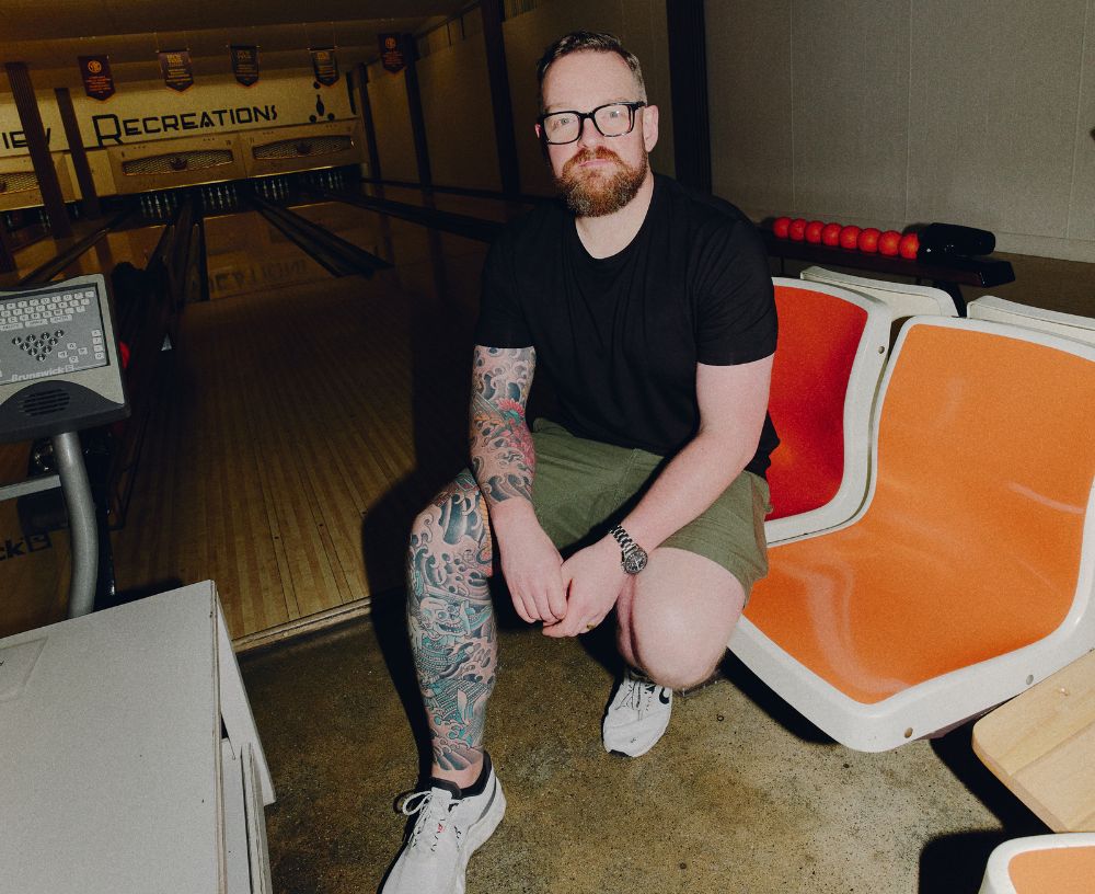 Man with tattoos and a beard sitting on an orange bench in a bowling alley.